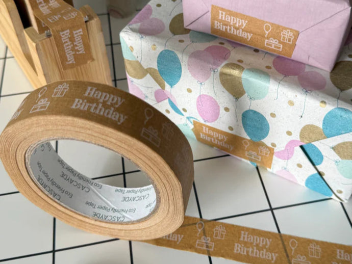 Roll of 'Happy Birthday' tape with wrapped gifts on a tiled floor